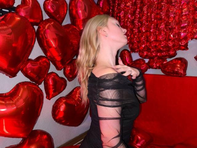 CrystalHope with blonde hair, wearing a black, sheer, long-sleeve dress, kissing a red heart balloon, surrounded by red heart balloons, her ass slightly visible.