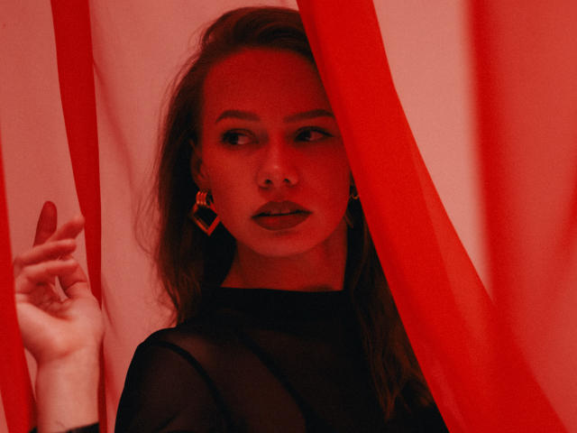 ShyySiren with red curtains, wearing black sheer top, holding curtain with left hand, looking to the side, wearing diamond-shaped earrings, red lighting, dark lipstick, long brown hair.