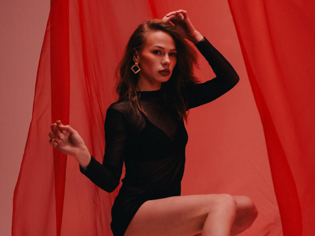 ShyySiren in a black sheer dress, showing her tits and ass, with red curtains, wearing gold earrings, standing with one leg up, and her hand in her hair.