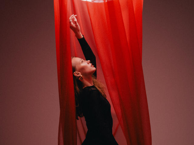 A photograph of ShyySiren standing with arms raised, partially obscured by red, translucent curtains. She wears a black long-sleeve dress, her blonde hair flowing down her back. The red curtains create a vertical frame around her. Her eyes are closed, and her head is tilted back, showcasing a relaxed, almost meditative posture. The red curtains and her black dress contrast sharply, emphasizing her profile. The soft, even lighting highlights her serene expression.
