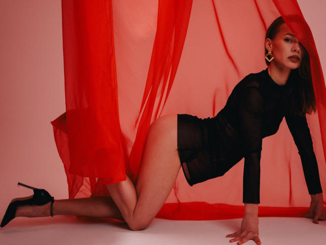 ShyySiren, a woman with fair skin and straight brown hair, kneels on a white floor wearing a sheer black long-sleeve top and black high heels. She's partially covered by a red sheer curtain that drapes behind her. She has a confident expression, dark lipstick, and large gold earrings. Her left hand is on the floor, and her right hand is behind her. The image uses bold red and black colors, emphasizing her toned, athletic physique. The red curtain adds a dynamic, flowing element to the composition.