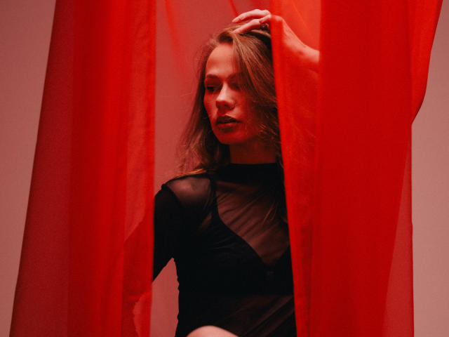 ShyySiren stands partially obscured by red, semi-transparent curtains. She has light brown, wavy hair and wears a black, sheer long-sleeve top. Her right hand is raised, gripping the curtain. Red light casts a warm glow, highlighting her facial features and the sheer texture of her top. The composition focuses on her expression and the contrast between the red curtains and black top. The image uses bold, red lighting to create a dramatic, intimate effect.