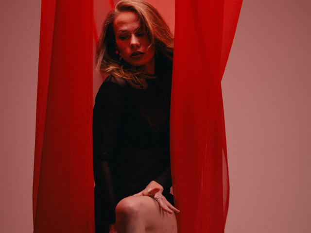 ShyySiren is partially hidden behind bright red curtains. She has light brown, wavy hair and a serious expression. She wears a black, sheer long-sleeve top, with one hand resting on her knee. Her other hand is out of frame. The red curtains are semi-transparent, creating a dramatic contrast with the dark top and her pale skin. The lighting is warm, highlighting her facial features and the red curtains. Her ring is noticeable on her right hand. The overall composition is bold and evocative, emphasizing the red curtains and ShyySiren's intense gaze.
