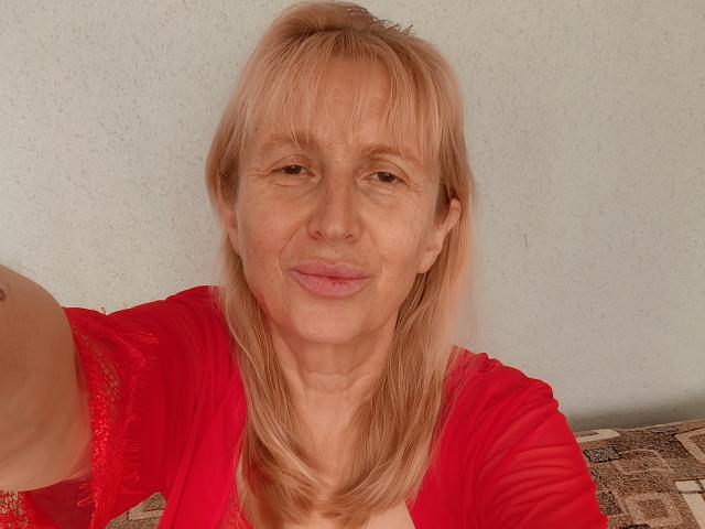 TrinaAdair with blonde hair, red shirt, and slight smile, showing her face and upper body, holding out arm for selfie. Her tits are small, and she has a tattoo.