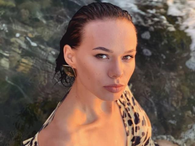 ShyySiren has wet, dark brown hair slicked back, and she's wearing large, gold, geometric earrings. She's looking directly at the camera with a neutral expression. She's wearing a leopard print top that's partially off her shoulders. The light is warm and casts a golden glow on her face. Her skin is smooth and she has a small mole on her right cheek. The background is blurred with green and brown textures. The overall composition is sharp with a focus on ShyySiren's face and upper torso. The image has a natural, candid feel.