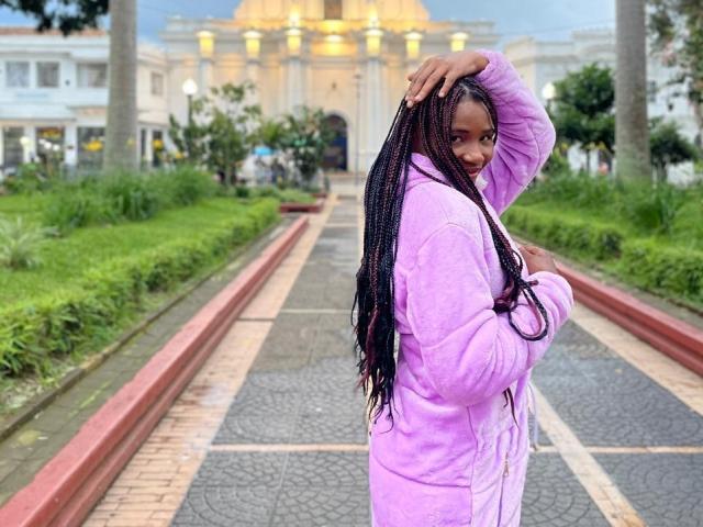 TenderBony with long black braids, wearing a pink fluffy jacket, standing on a brick pathway, hand on head, looking over shoulder, in a sunny, urban park setting.