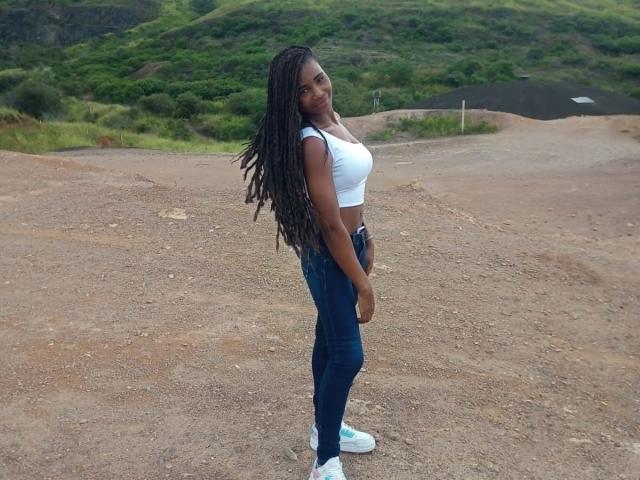 TenderBony with long black dreads, white crop top, showing her small tits, wearing blue jeans and white sneakers, standing on a dirt path, smiling.