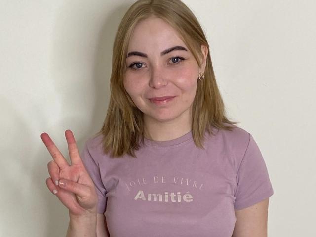 ShaquitaRasul with blonde hair, light skin, small tits, wearing a purple shirt, smiling, making a peace sign with her right hand, against a plain white background.