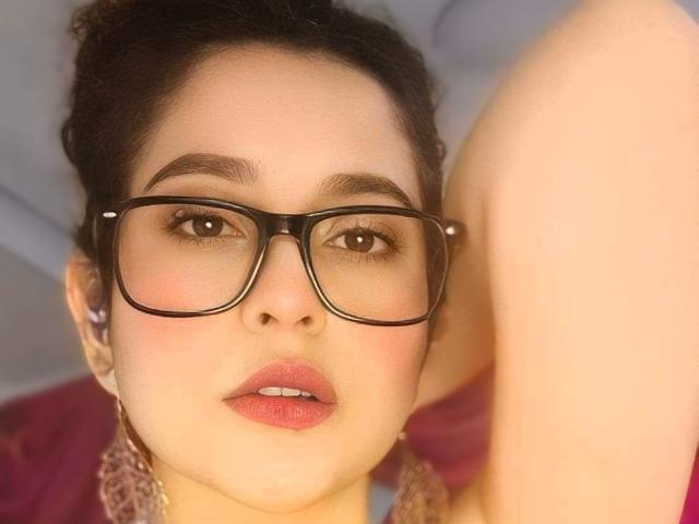 IrinaSweett with black-framed glasses, pink lipstick, and brown eyes, looking at the camera with a slight pout, arm raised, and dark hair in a messy bun.