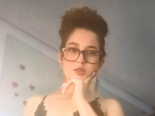 IrinaSweett with dark curly hair, glasses, and black lace lingerie, hand near mouth, looking at camera, pale skin, soft light, slightly blurred, sexy, subtle makeup, confident expression.