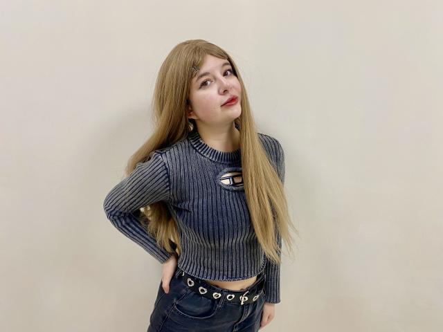 Reginia with long blonde hair, wearing a ribbed blue crop top and dark jeans with heart-shaped belt buttons, hands on hips, looking up with a slightly pouty expression.