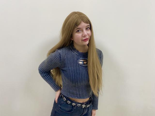 Reginia has long blonde hair, a ribbed blue crop top, and blue jeans with a belt. She's smiling, hands on hips, looking to the side. Her tits are small.