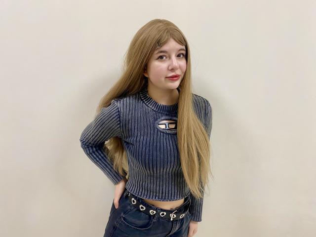 Reginia with long blonde hair, blue ribbed crop top, and dark jeans, hands on hips, slight smile, looking to the side, standing against a plain wall.