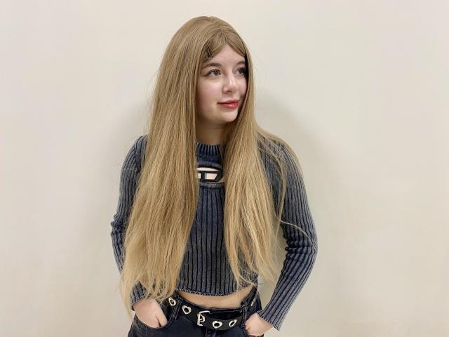 Reginia with long blonde hair, wearing a dark ribbed crop top and black belt, hands in pockets, standing against a plain wall, smiling slightly.