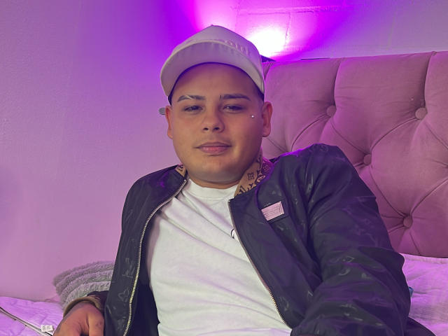 AaronKingg, chubby, wearing a white cap, black jacket, and white tee, sitting on a bed with a pink-purple light behind him, looking slightly annoyed with his tongue out.