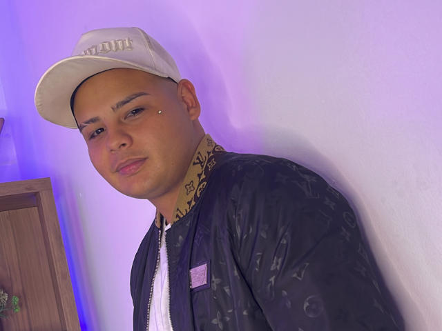 AaronKingg, tan-skinned, wearing a white cap, black patterned jacket, and a white shirt, leaning against a white wall with a subtle purple light. Small piercing on right cheek.