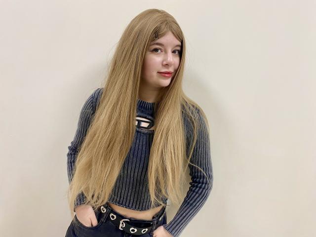 Reginia with long blonde wig, blue ribbed crop top, and black belt, hands in pockets, smiling slightly, pale skin, red lipstick, standing against plain wall.