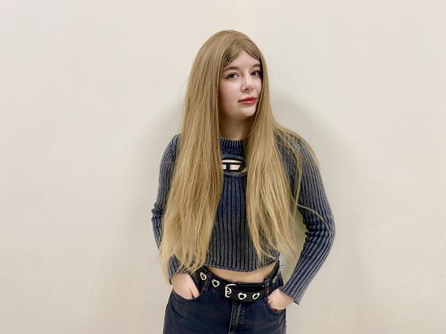 Reginia with long blonde hair, blue striped crop top, hands in black jeans with belt, standing, neutral expression, light skin, red lipstick, slim build, casual style.