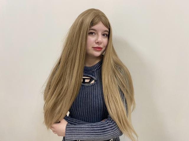 Reginia with long, straight blonde wig, wearing a blue ribbed sweater with a black emblem, standing against a plain wall, hands in front, slightly smiling with pink lips.