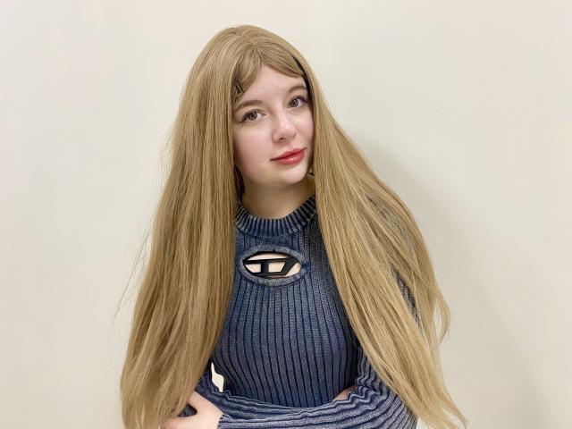 Reginia with long blonde hair, blue ribbed sweater, and cute smile, has her tits slightly visible through the sweater's cutout, looking at the camera.