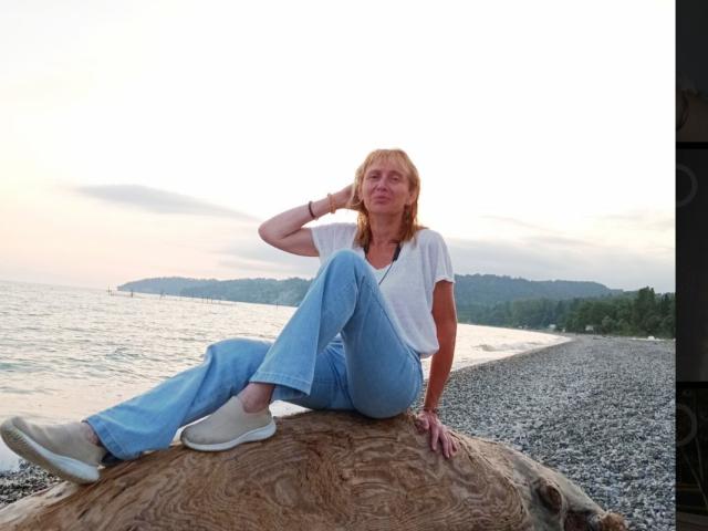 TrinaAdair, middle-aged woman with red hair, sits on a log, wearing blue jeans and white shoes, white tee, hand on head, looking relaxed, by the pebble beach.