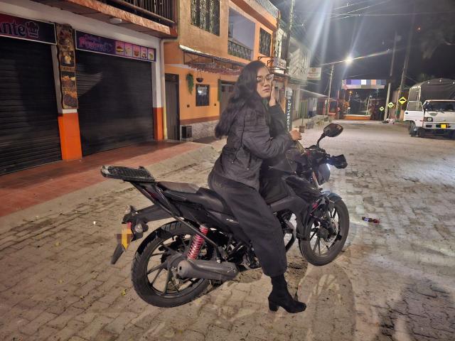 SofiaMonseur with long black hair, puffy jacket, black pants, and black boots, sitting on a black motorcycle at night, holding the handlebars, on a cobblestone street.