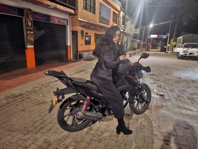 SofiaMonseur with long dark hair, wearing a puffy jacket and black pants, sits on a black motorcycle at night, wearing black boots, on a cobblestone street.
