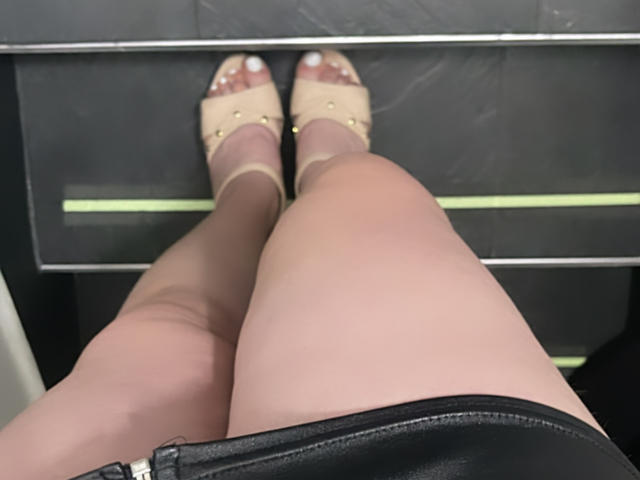 JennyLawry's legs in sheer pantyhose, wearing beige peep-toe heels with gold studs, sitting on a black surface, black leather skirt visible at the bottom.