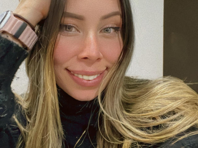 JennyLawry has blonde and brown hair, a pink watch, black turtleneck, and is smiling with her hand in her hair. Her tits are not visible. She has a cute face.