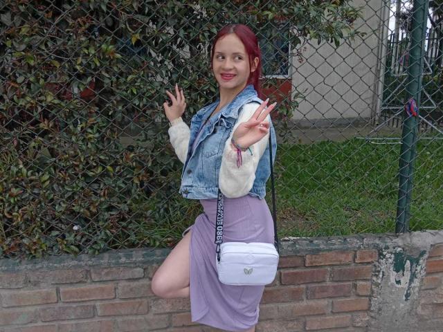 LesleyGray with red hair, denim vest, white long-sleeve shirt, purple skirt, white purse, showing off tits, and giving a peace sign, standing against a brick wall.