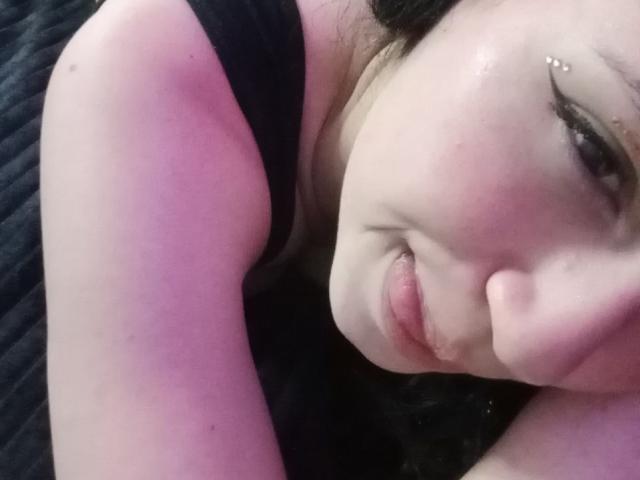 A SofiaFaces tem pele clara, delineador preto e um piercing no nariz. Ela está de língua para fora, com o braço levantado, vestindo uma blusa preta, em um selfie com iluminação rosa..