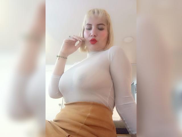  GiuliaAddams with blonde bob, red lipstick, white sheer top showing her tits, and tan skirt, holding her cheek, wearing a bracelet, in a bright room.