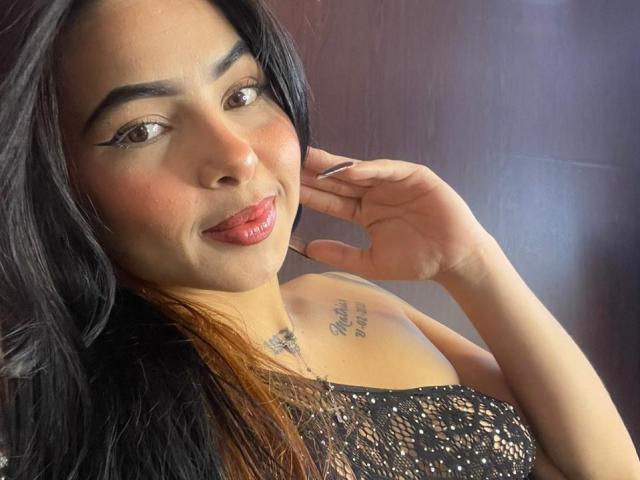 MistressAsshley with long black hair, brown eyes, and glossy pink lips, wearing a black lace top, has a tattoo on her chest, and is touching her ear.