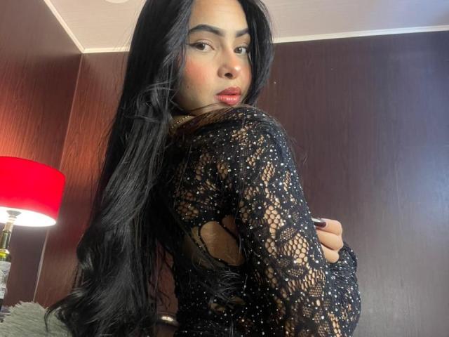 MistressAsshley, con lunghi capelli neri, indossa una camicetta di pizzo nero, e guarda oltre la spalla, mostrando la schiena e i glutei. In secondo piano, una lampada rossa..