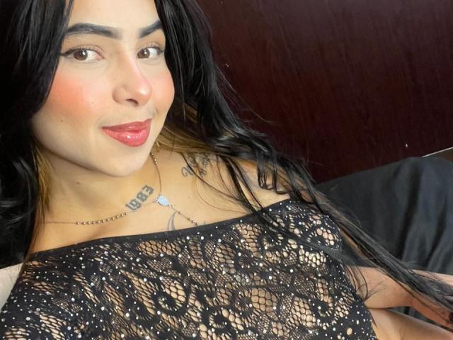 MistressAsshley, con cabello negro largo, labios rosados y tatuajes, vestida con una blusa negra de encaje que deja ver sus pechos, sonriendo a la cámara. Sus pezones son visibles a través del encaje..