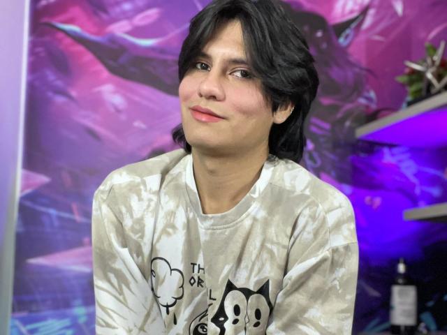 AlexxNorth with medium-length black hair, wearing a white and gray tie-dye shirt, smiling slightly, sitting with arms at sides, in a purple-lit room.