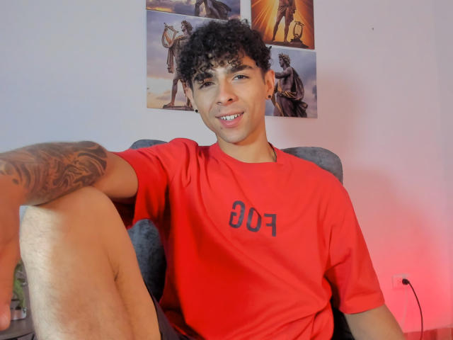 AlexZT with curly black hair, tan skin, tattooed arm, and red shirt, sitting, smiling, knees up, in a gray chair, red light in background.