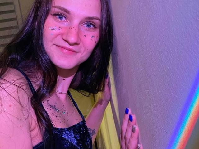 ErrosAy with long brown hair, blue eyes, and sparkly freckles, smiling, wearing a black sequined top, blue nails, touching yellow wall, neon rainbow on right.