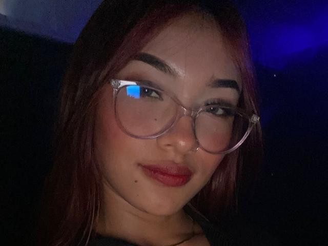 SashaHans has red hair, glasses, and is wearing red lipstick with a small mole on her right cheek, looking slightly to the left with a subtle smile.