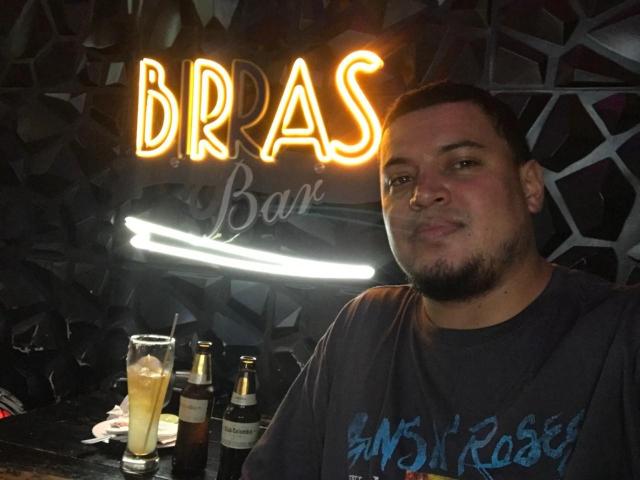 AragonWel, a chubby dude with short dark hair and a beard, wearing a gray shirt with blue text, sits by a bar with drinks and a neon sign.