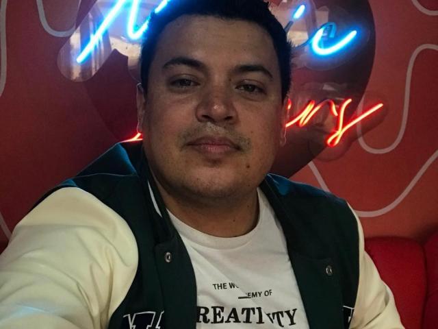 AronWel, a chubby dude with short black hair, wearing a green and white varsity jacket, looking at the camera with a slight smile, neon lights in the background.