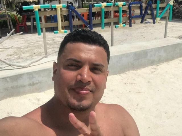 AronWel, shirtless, has a tan, muscular build, short black hair, and a goatee. He's making a peace sign with a smile, standing on a sandy beach.
