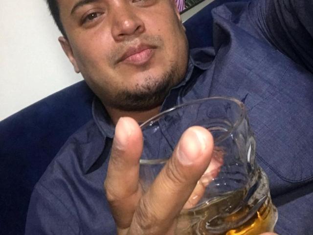 AragonWel, a chubby man with short black hair and a goatee, holding a glass of whiskey, puffs out his cheek with a smug expression, wearing a blue shirt.