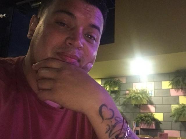 AragonWel with short black hair, red shirt, and heart tattoo on right wrist, pouting with hand on chin, under purple lighting. His wrist tattoo is above his ass.