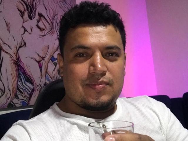 AragonWel, a chubby dude with short black hair and a goatee, wearing a white shirt, holding a glass, and giving a slight smirk. Anime-style wall art in background.
