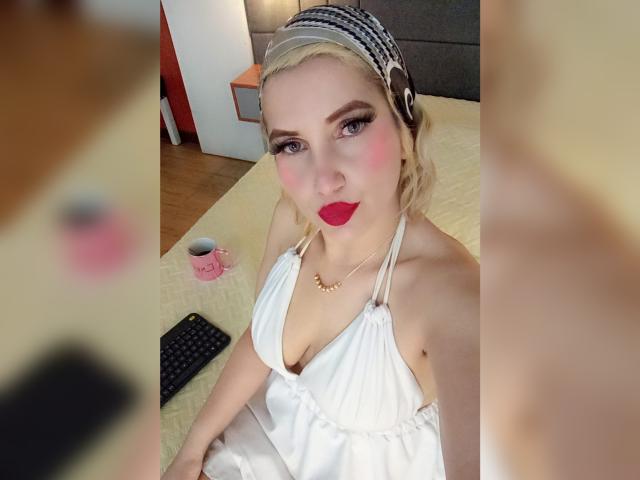  GiuliaAddams with blonde hair, red lipstick, white halter dress, and headphones, sitting on bed, holding pink mug, remote, and wooden floor in background. Boobs visible.