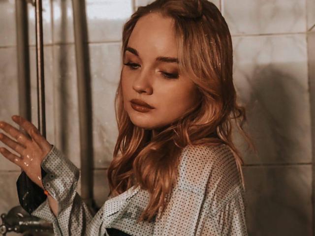 ShawtyMiaAndJack with wavy blonde hair, wearing a polka-dot shirt, standing in a shower, water on her hand, looking down with a serious expression, sexy and sultry.