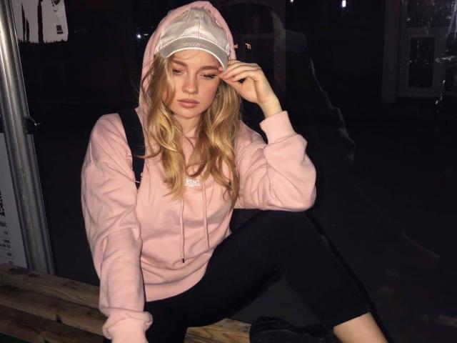 ShawtyMiaAndJack with long blonde hair, pink hoodie, black pants, and white cap, sitting on a wooden bench at night, touching her face, looking serious, with a backpack.