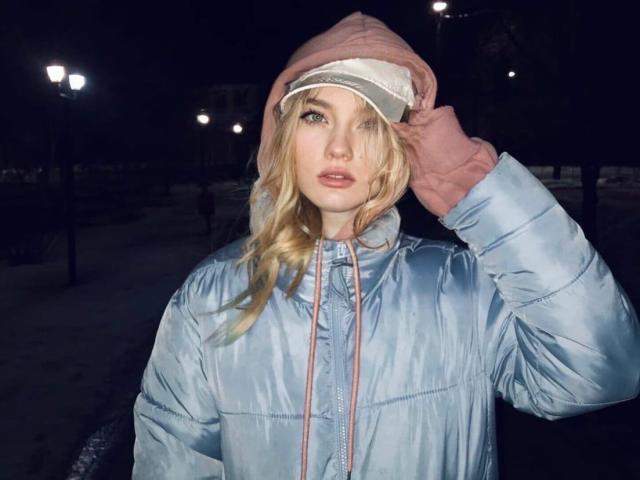 ShawtyMiaAndJack with blonde hair, blue eyes, wearing a pink hood, light blue puffy jacket, pink gloves, holding hat, night, street lamps in background.