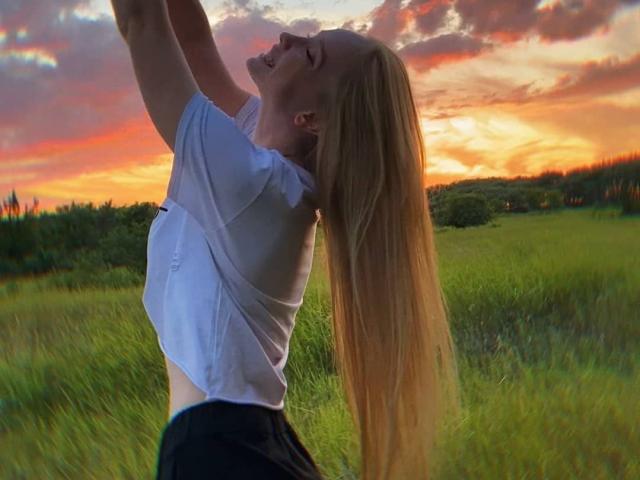 ShawtyMiaAndJack with long blonde hair, white shirt, and black pants, arms raised, head tilted back, smiling at a colorful sunset, grassy field in the background.
